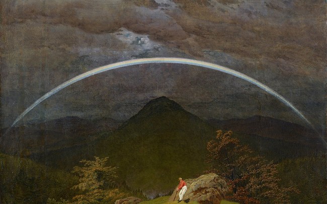 A rainbow arching across a dark, cloudy sky over a mountain, with a lone traveler resting on a rock below