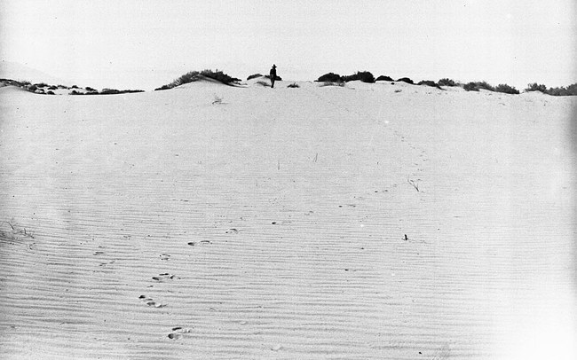 A lone man in a hat walking across wind-swept desert sand, his footprints trailing behind him